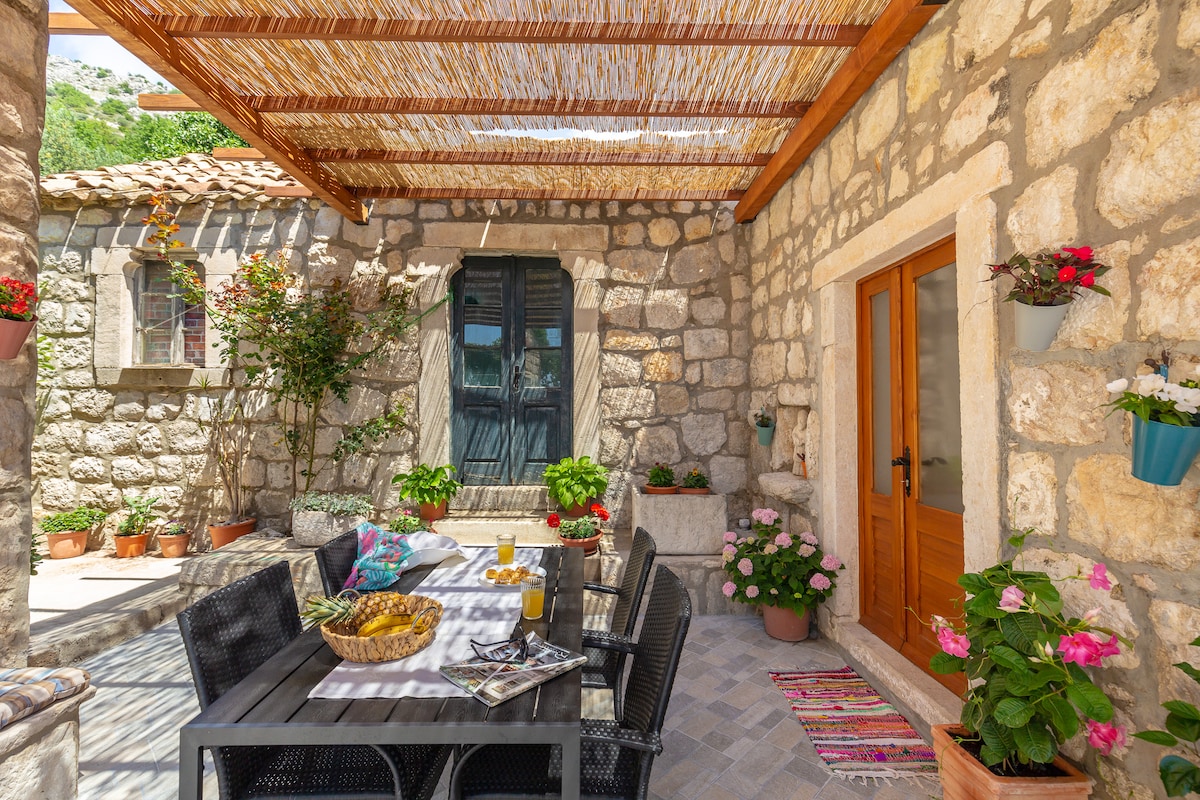 A shaded terrace is displayed, featuring a dining table with comfortable chairs and a selection of fresh fruits. Potted flowers add color to the stone walls, while a wooden entrance door and large window offer glimpses of the cottage. The atmosphere is serene and inviting.