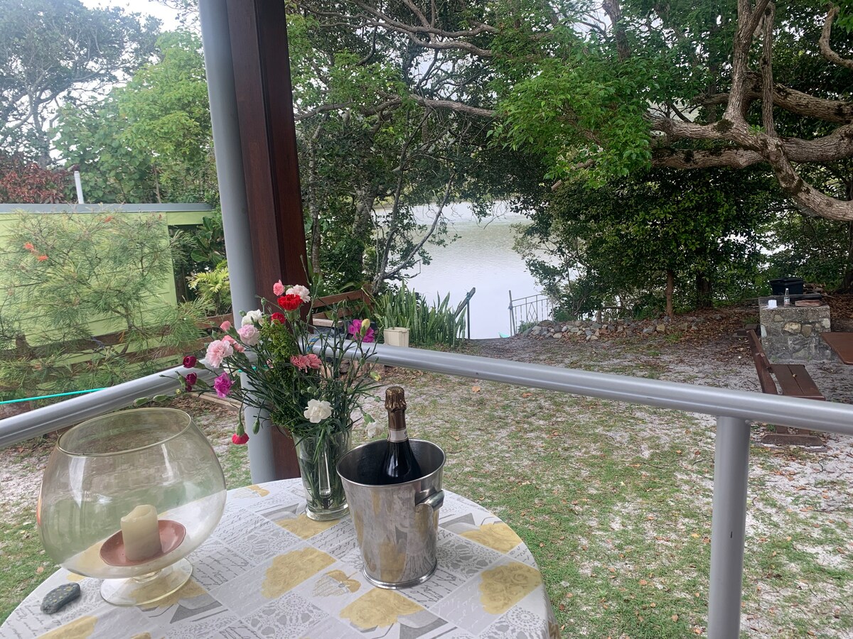 A covered outdoor space features a round table set with a floral arrangement, a candle, and a bottle of champagne in an ice bucket. The view overlooks Cudgera Creek, surrounded by lush greenery, providing a peaceful setting for relaxation.