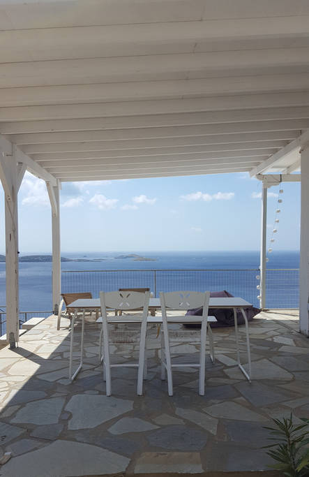 An outdoor terrace features a dining table surrounded by white chairs, set against a backdrop of the Aegean Sea. The clear blue sky and distant islands create a serene view, while the stone flooring adds a natural element to the space.