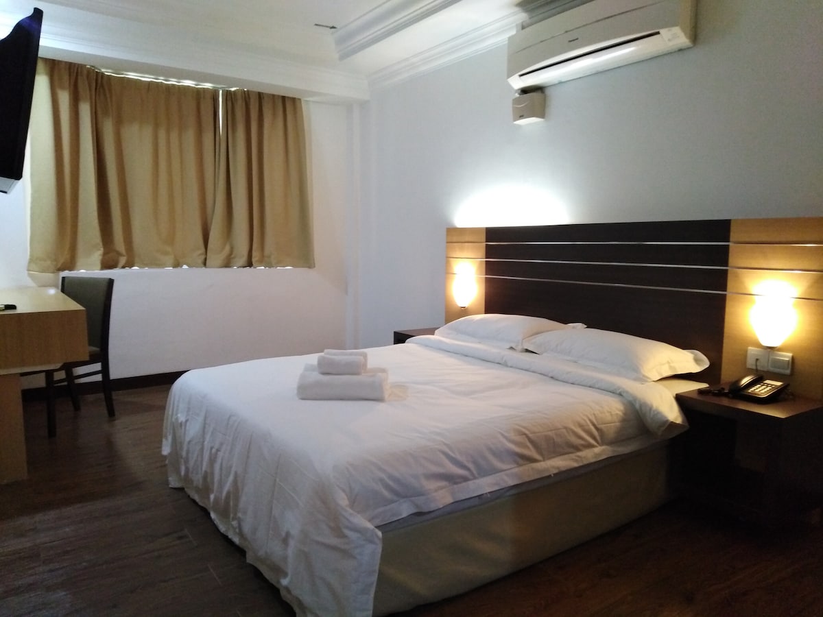 A simple room features a spacious bed with neatly arranged white linens and towels. A wooden desk and chair are positioned beside the window adorned with light brown curtains. Soft lighting creates an inviting atmosphere, while an air conditioning unit and a television are mounted on the wall.