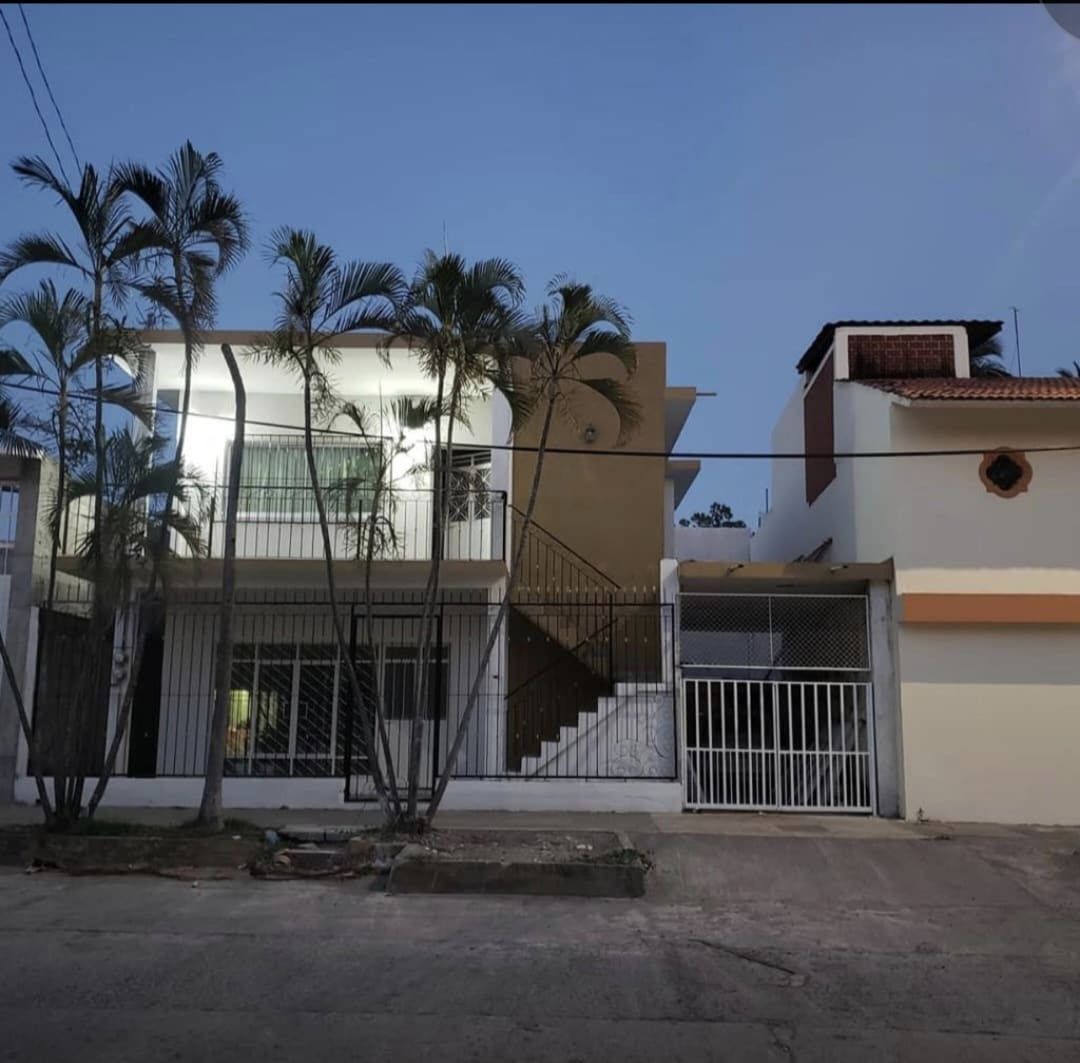 A two-story building is positioned amidst palm trees, featuring a welcoming entrance with a staircase leading to a balcony. The exterior walls are painted in neutral tones, with varied architectural styles evident in the neighboring structures.