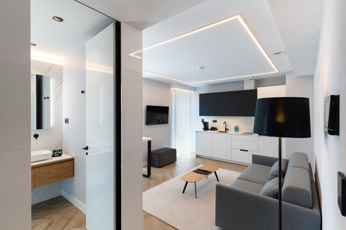 The open living area reveals a modern sofa facing a sleek coffee table. A television is mounted on the wall opposite a stylish kitchen featuring cabinetry and appliances. Natural light enters through floor-to-ceiling windows, enhancing the minimalist design with clean lines.