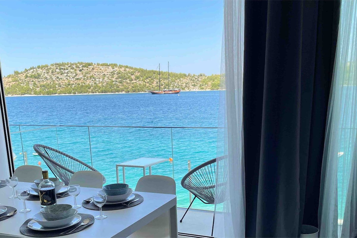 A dining area is visible, set with elegant tableware and overlooks the vibrant blue sea. The glass doors provide a panoramic view of the tranquil waters and the lush hillside beyond, with a sailboat gently anchored in the distance.