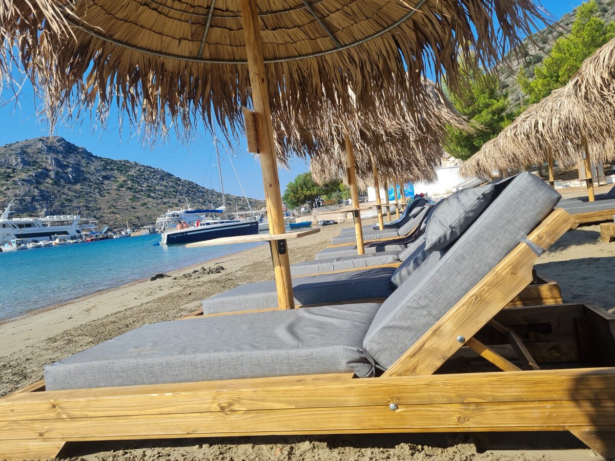 Lounge chairs are positioned under thatched umbrellas along a sandy beach. The sea glistens nearby, with boats visible in the water. The landscape includes a distant mountain, creating a serene coastal atmosphere.