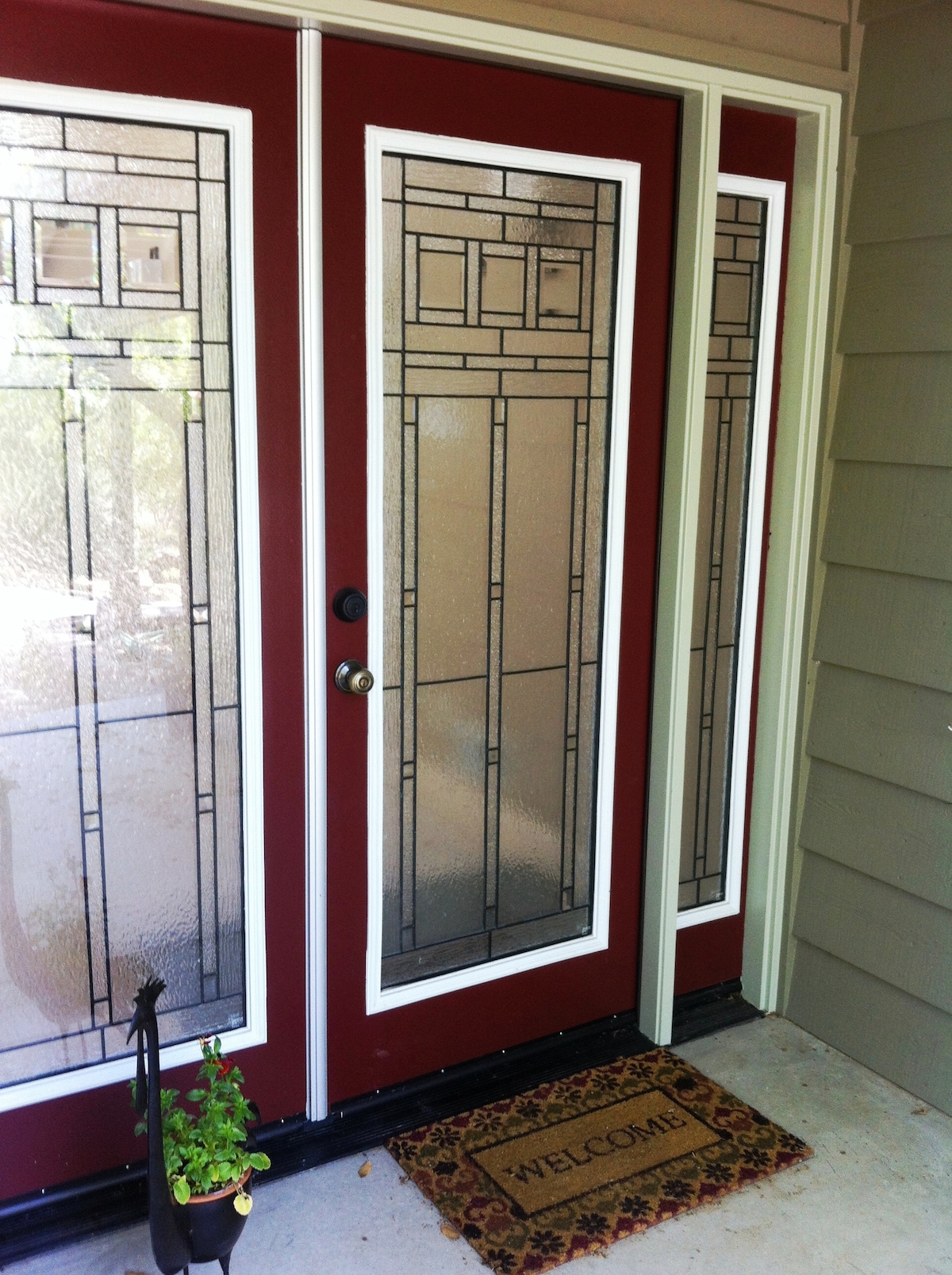 The entrance showcases a pair of elegant glass-paneled doors with decorative designs, framed in rich red. A small potted plant is positioned beside the doors, which feature a welcome mat inviting guests into the space.