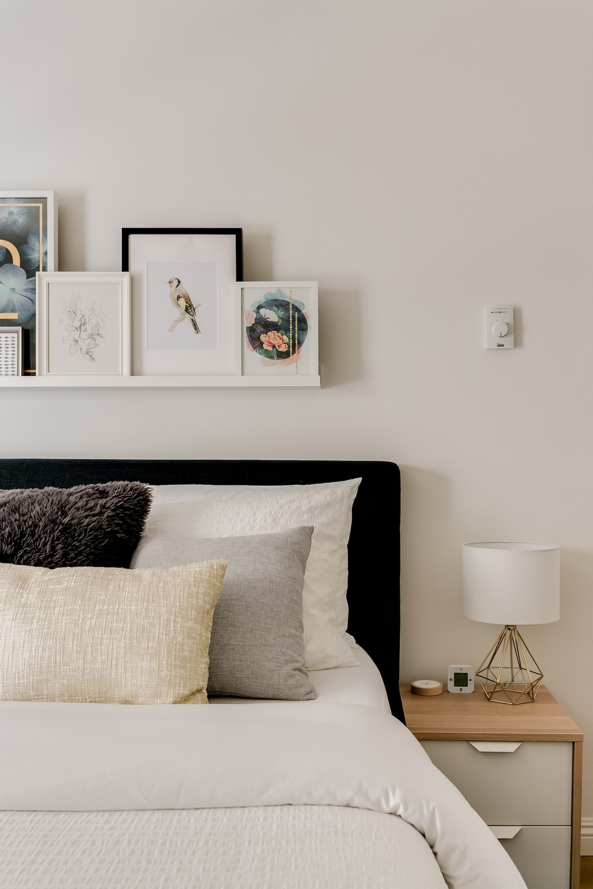 A neatly made bed is adorned with a variety of decorative pillows, resting against a simple headboard. Above the bed, an art display features framed botanical prints and images of birds, while a nightstand holds a lamp and small electronic device.