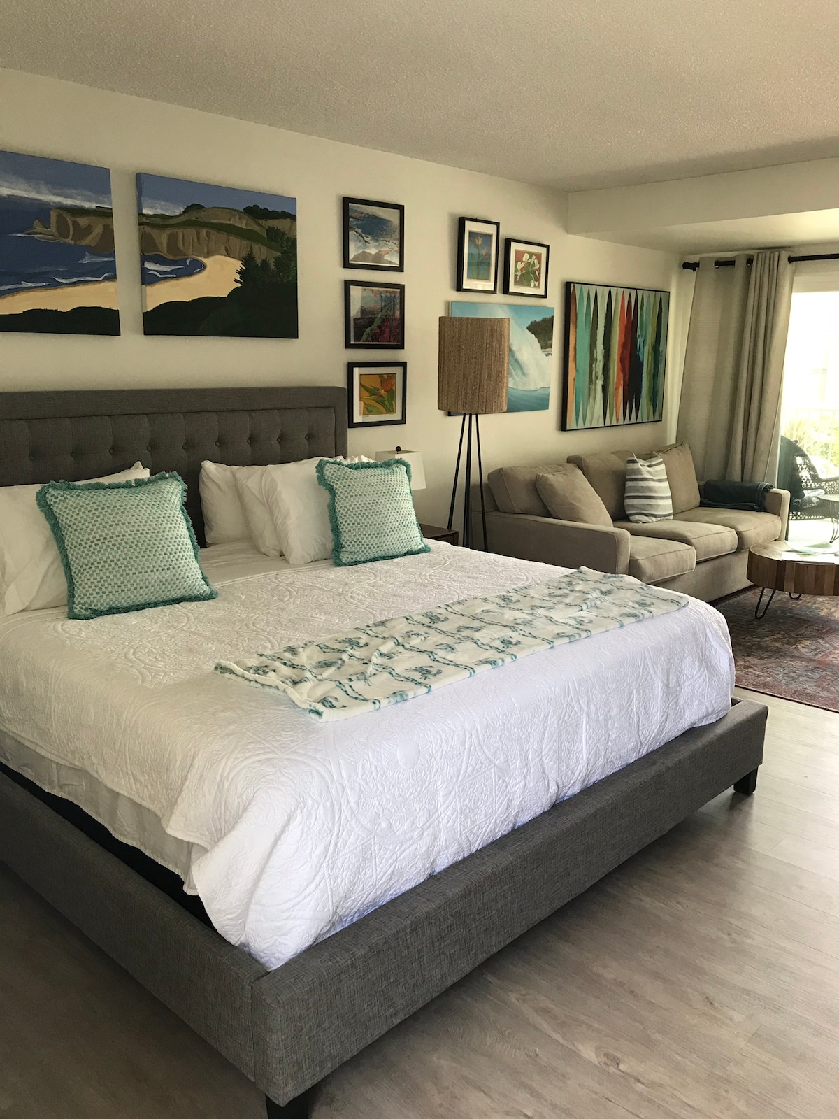 A spacious sleeping area features a king size bed with white linens and decorative pillows. The room is adorned with colorful artwork on the walls. A comfortable seating area includes a couch, accented by natural light streaming through large windows with soft drapes.
