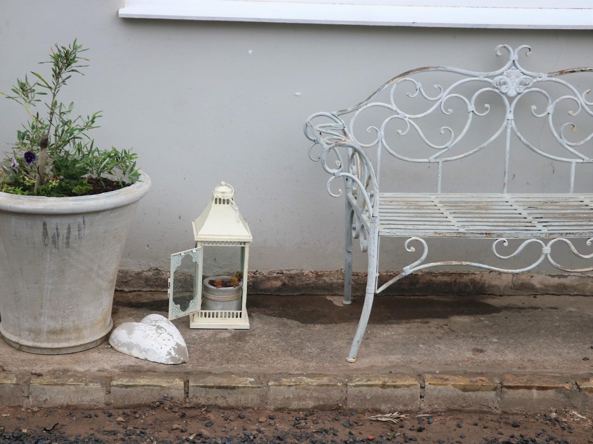 A white, vintage-style wrought iron bench is situated alongside a large planter filled with greenery. A decorative lantern, positioned on the ground, adds charm to the outdoor space. The patio's stone pathway is visible, leading to the inviting entrance.