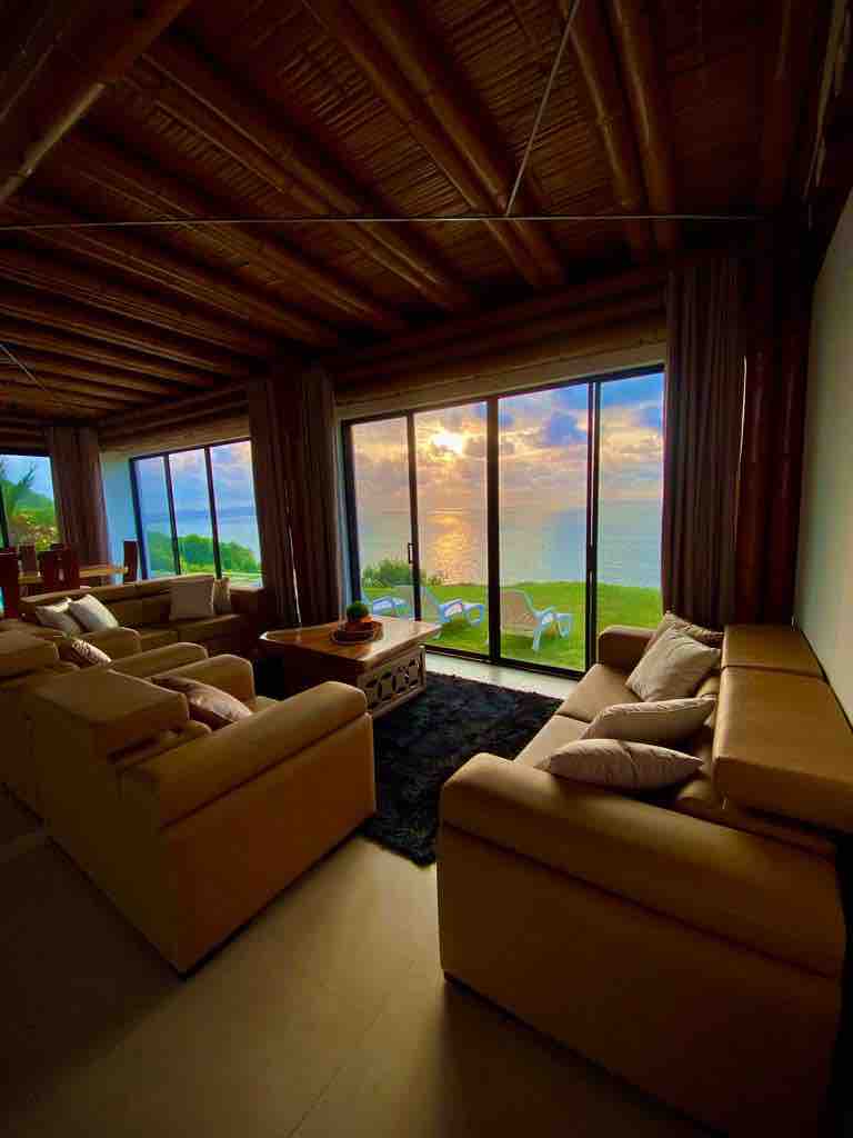 A spacious living area features light-colored sofas arranged around a central wooden coffee table. Large glass doors provide a view of the sunset over the ocean, with the soft reflection of colors visible on the water. Natural light fills the room, enhancing the tranquil ambiance.
