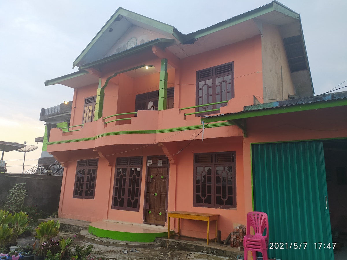 A two-story building features vibrant peach-colored walls with green accents. The front porch is supported by columns, with a wooden door at the center. Large windows with grills provide ample light, while a table and a pink chair are situated outside, complemented by surrounding greenery.
