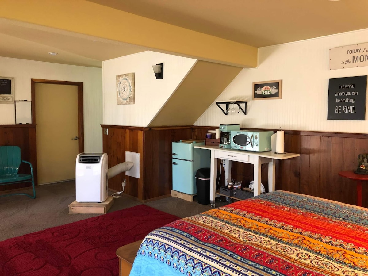 The room features a vintage-style kitchenette with a turquoise refrigerator and microwave. A spacious bed, adorned with a brightly colored blanket, complements the wooden accents. A comfortable chair and portable air conditioner are positioned nearby, with a door leading to the rest of the apartment.