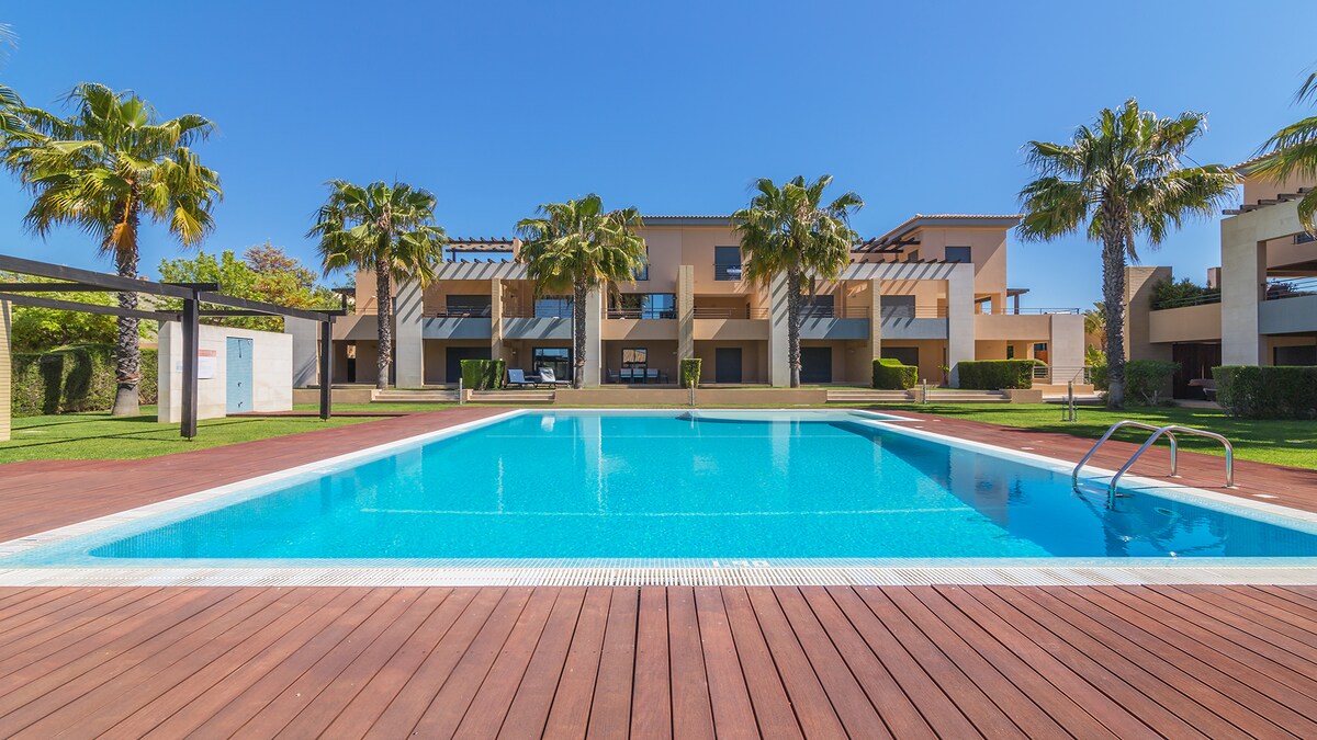 A large swimming pool is surrounded by a wooden deck, with several palm trees providing greenery in the background. The two-story buildings, featuring modern architectural elements, are visible beyond the pool area, set against a clear blue sky.