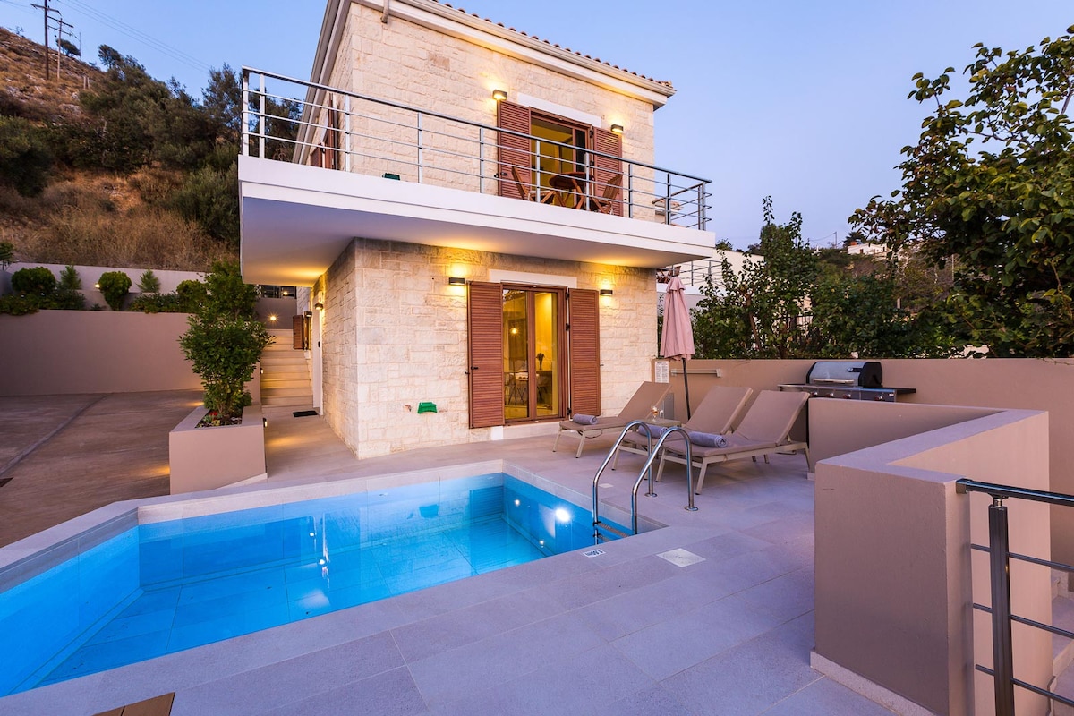 A modern villa exterior is presented, featuring a private pool with clear blue water and sun-loungers nearby. The stone façade is complemented by wooden shutters, and an outdoor dining area with a barbecue is visible, all set against a backdrop of lush greenery.