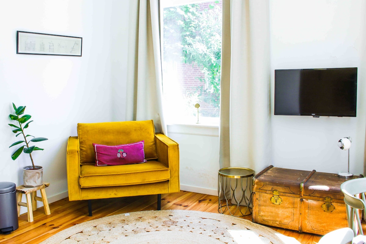 A bright corner of the living area features a vibrant yellow armchair with a pink accent pillow. A small side table and a potted plant are positioned nearby, alongside a television mounted on the wall. Natural light streams through a window framed by light curtains.