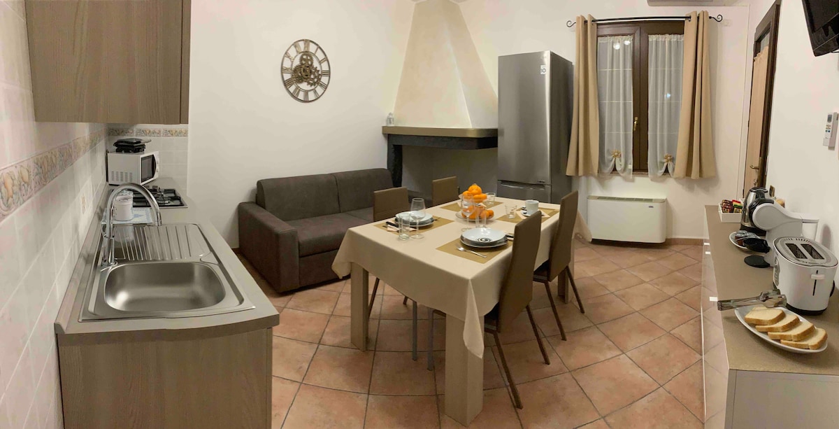 A functional kitchen is shown with a dining table set for four, complemented by a cozy sofa in the corner. Appliances include a refrigerator and a variety of cooking tools. Natural light enters through the window, enhancing the warm tones of the tiled floor.