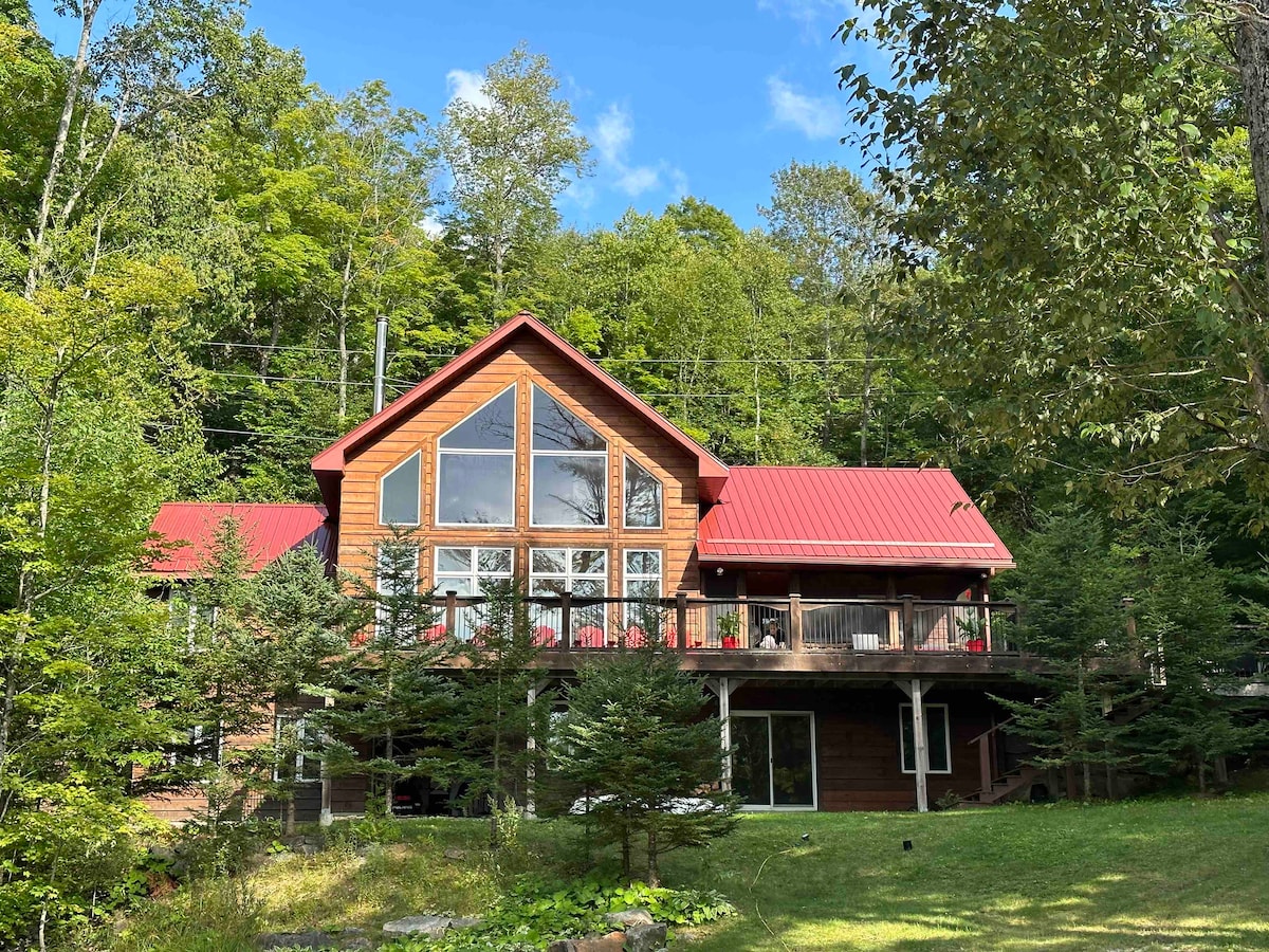 The exterior of a large lakefront cottage is showcased, featuring a striking red metal roof and multiple tall windows. Surrounded by lush greenery, the building presents a spacious deck and a well-maintained landscape, inviting outdoor relaxation.