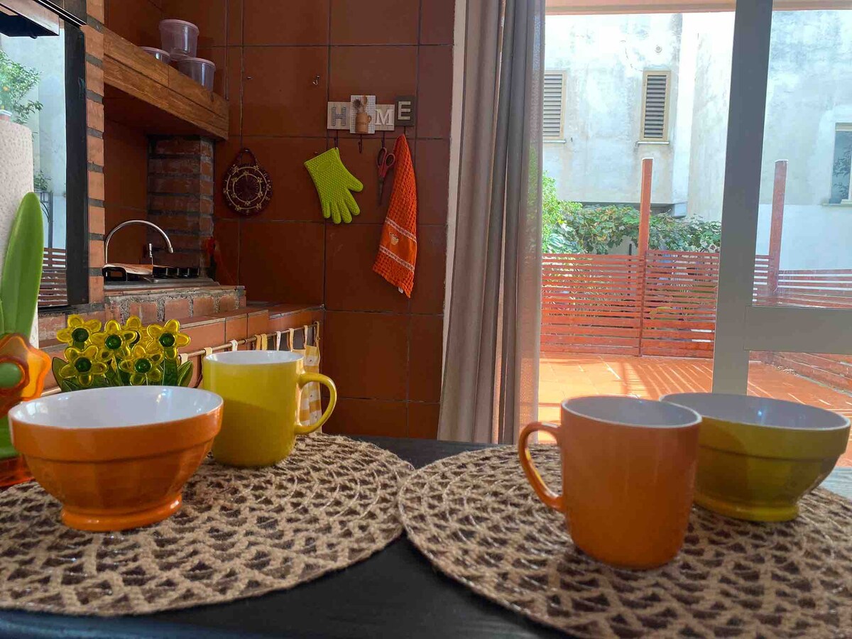 The image depicts a kitchen area featuring a rustic brick wall with cooking utensils hanging. On the table, four colorful cups and bowls are arranged on round woven placemats. Sunlight enters through a window, illuminating a small outdoor space visible in the background.