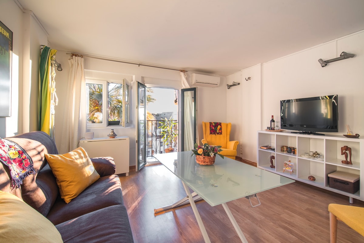 A well-lit living area features a comfortable brown sofa adorned with decorative pillows and a glass dining table at its center. Large windows allow natural light and provide access to a small balcony. A bright yellow armchair and a television are positioned nearby.