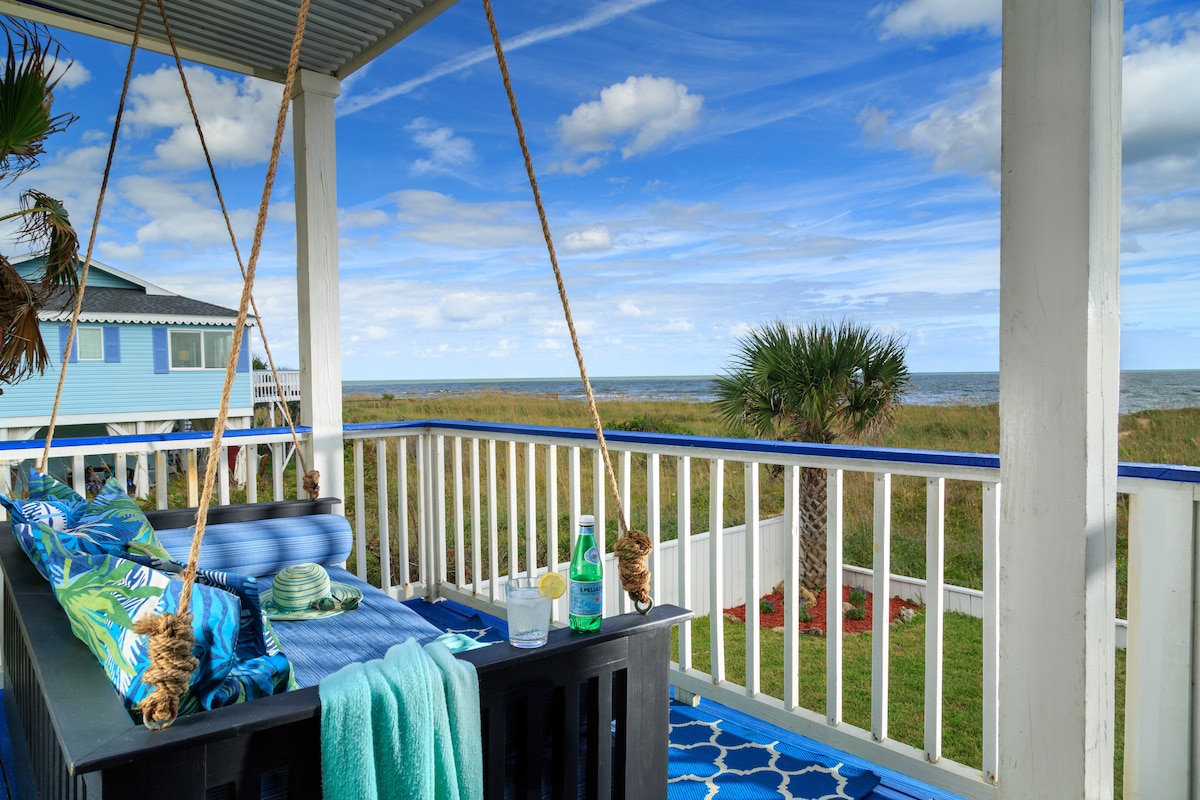 An outdoor porch features a comfortable swinging bed adorned with colorful cushions and blankets. The surrounding railing offers unobstructed views of the ocean and nearby palm trees. A vibrant blue patterned rug complements the tropical setting, enhancing the relaxing atmosphere of the space.