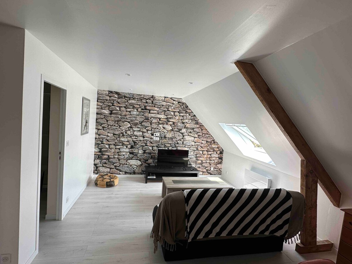 A welcoming living area is defined by a natural stone accent wall and sloped ceilings. A comfortable sofa with a black and white striped throw faces a low coffee table. A long window allows light to fill the space, while a small pet bed is situated nearby.