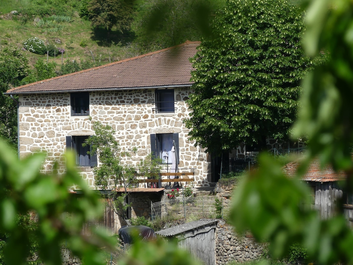The exterior of the gîte is showcased, featuring a traditional stone façade and large windows. A welcoming porch area with wooden railings and a neat garden is visible. Lush greenery surrounds the building, enhancing the natural setting in the countryside.