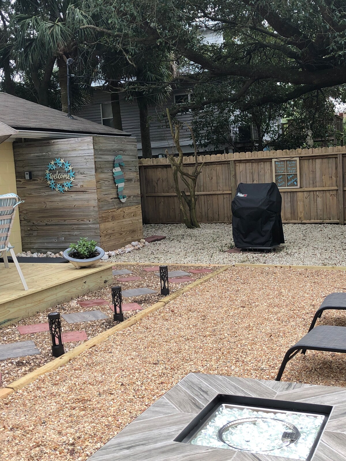 A private outdoor space is enclosed by an 8-foot wooden fence, featuring a gravel yard with decorative pathways. A gas grill is covered in the corner, and comfortable lounge chairs are positioned nearby, creating an inviting area for relaxation and outdoor meals.
