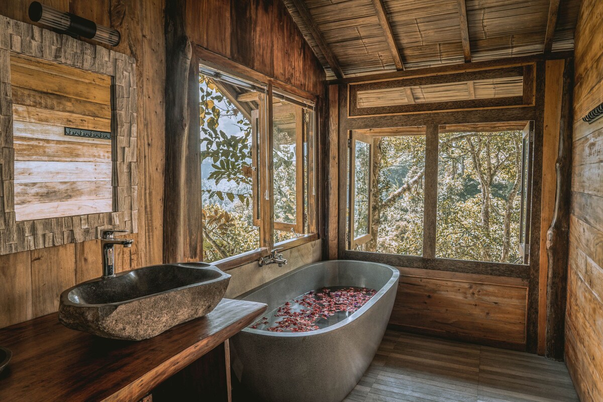 A spacious bathroom features large windows that provide a view of the surrounding nature. A freestanding bathtub, filled with rose petals, is complemented by a stone sink and wooden accents, creating a serene and natural atmosphere.