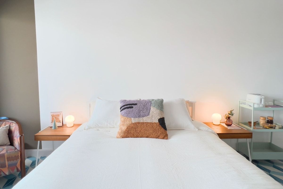 A spacious bed is positioned against a white wall, accompanied by soft lighting from bedside lamps. A decorative pillow adds a touch of color, while a small side table holds a plant and a few personal items.