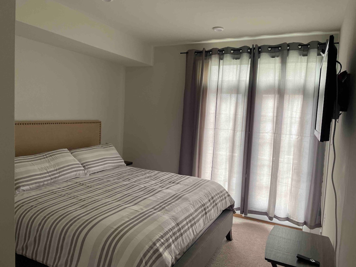 The bedroom features a spacious bed with a striped comforter and neutral-toned headboard. Soft curtains gracefully drape over the window, allowing natural light to softly filter in. A small table is positioned beside the bed, complemented by a wall-mounted television.