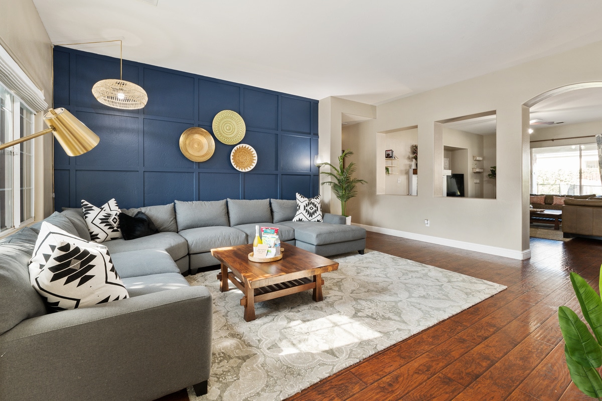 A spacious living area is highlighted by a large sectional sofa arranged around a coffee table. A textured area rug lays beneath, complementing the warm wood floor. Decorative wall plates are displayed on a blue accent wall, with abundant natural light highlighting the space.