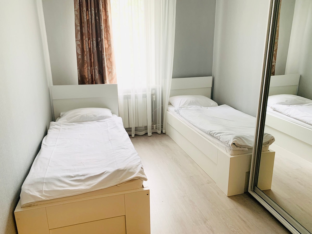 A bright sleeping area features two single beds arranged near a large mirrored wardrobe. Soft white bedding is used on both beds. Natural light filters in through sheer curtains, illuminating the light wood flooring and the pale grey walls.