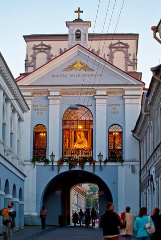 An archway leads to a historic building featuring intricate architectural details. Stained glass windows display an image of religious significance, framed by ornamental decorations. People can be seen walking through the arch, contributing to the location's lively atmosphere.