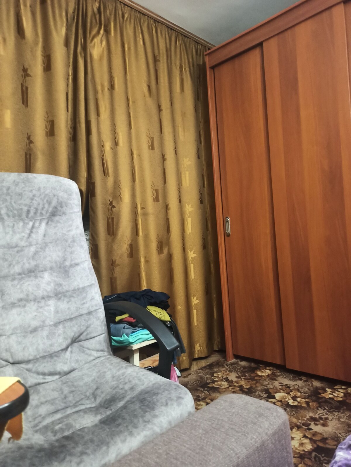 A textured gray armchair is positioned next to a wooden wardrobe. Golden curtains drape along the wall, contrasting with the warm tones of the furniture. A small shelf with colorful clothing is visible, adding a touch of vibrancy to the cozy room.