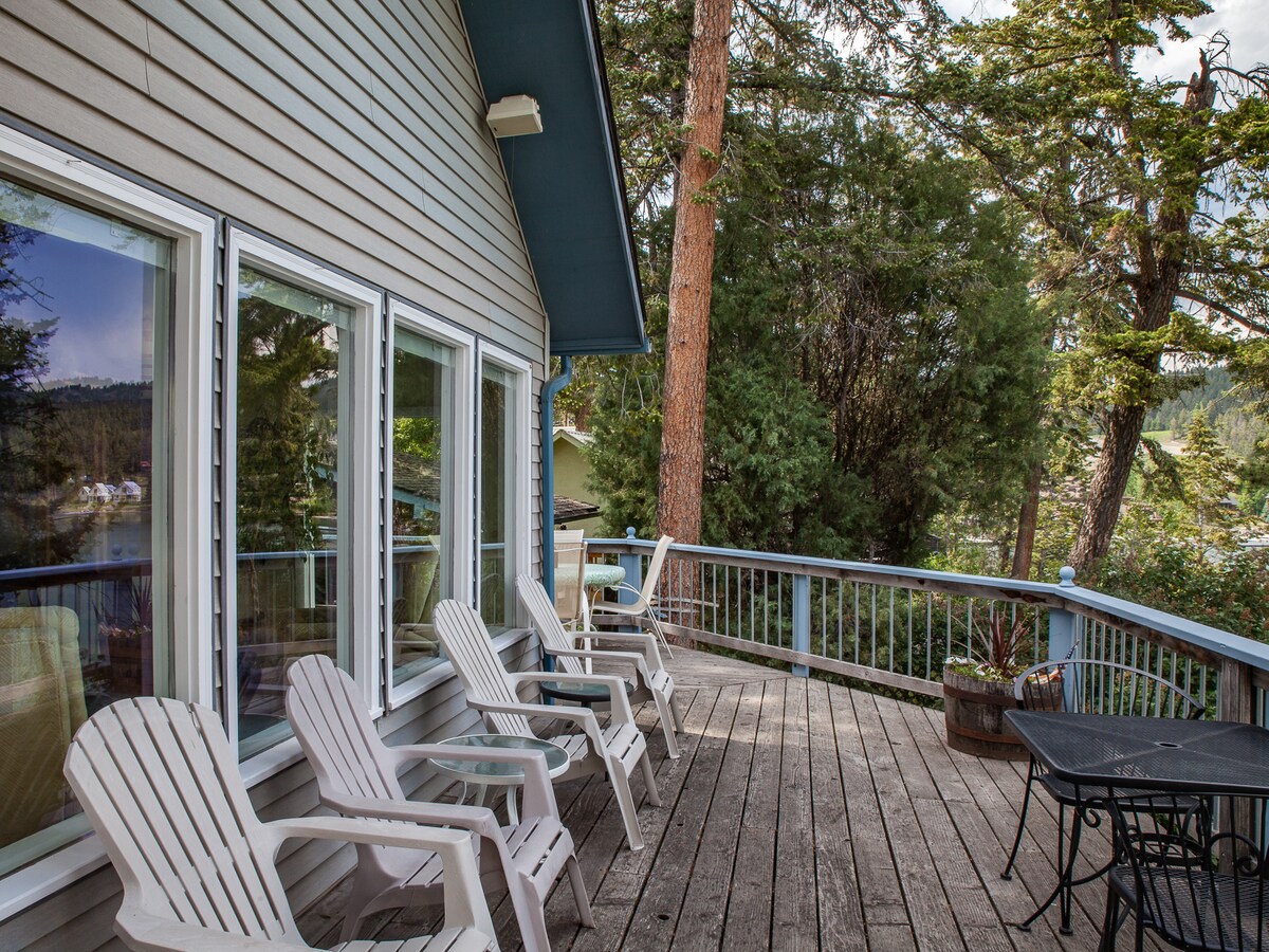 A spacious wooden deck is presented, featuring several white lounge chairs arranged for relaxation. The surrounding trees and nearby views create a serene outdoor setting. Large windows of the cabin reflect natural light and provide glimpses of the scenic landscape.