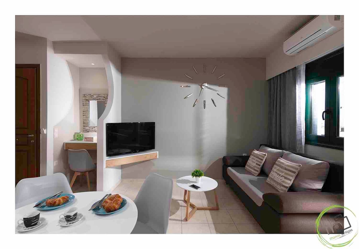 A cozy living area features natural light illuminating a welcoming space with a comfortable sofa. A modern television is mounted on the wall, while a round coffee table is set with crockery. Light curtains frame the windows, enhancing the overall ambience.