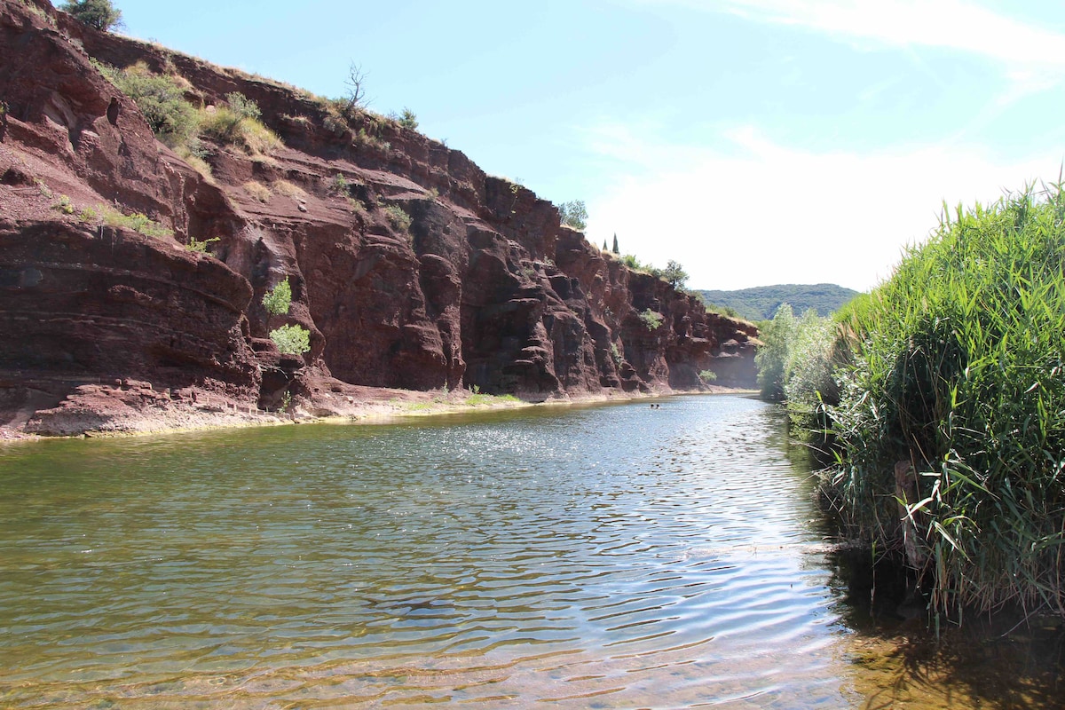 Clear water reflects the blue sky, surrounded by red rocky cliffs and greenery. The area shows a tranquil riverbank, where gentle slopes of vegetation lead to the calm surface, inviting moments of relaxation and exploration.