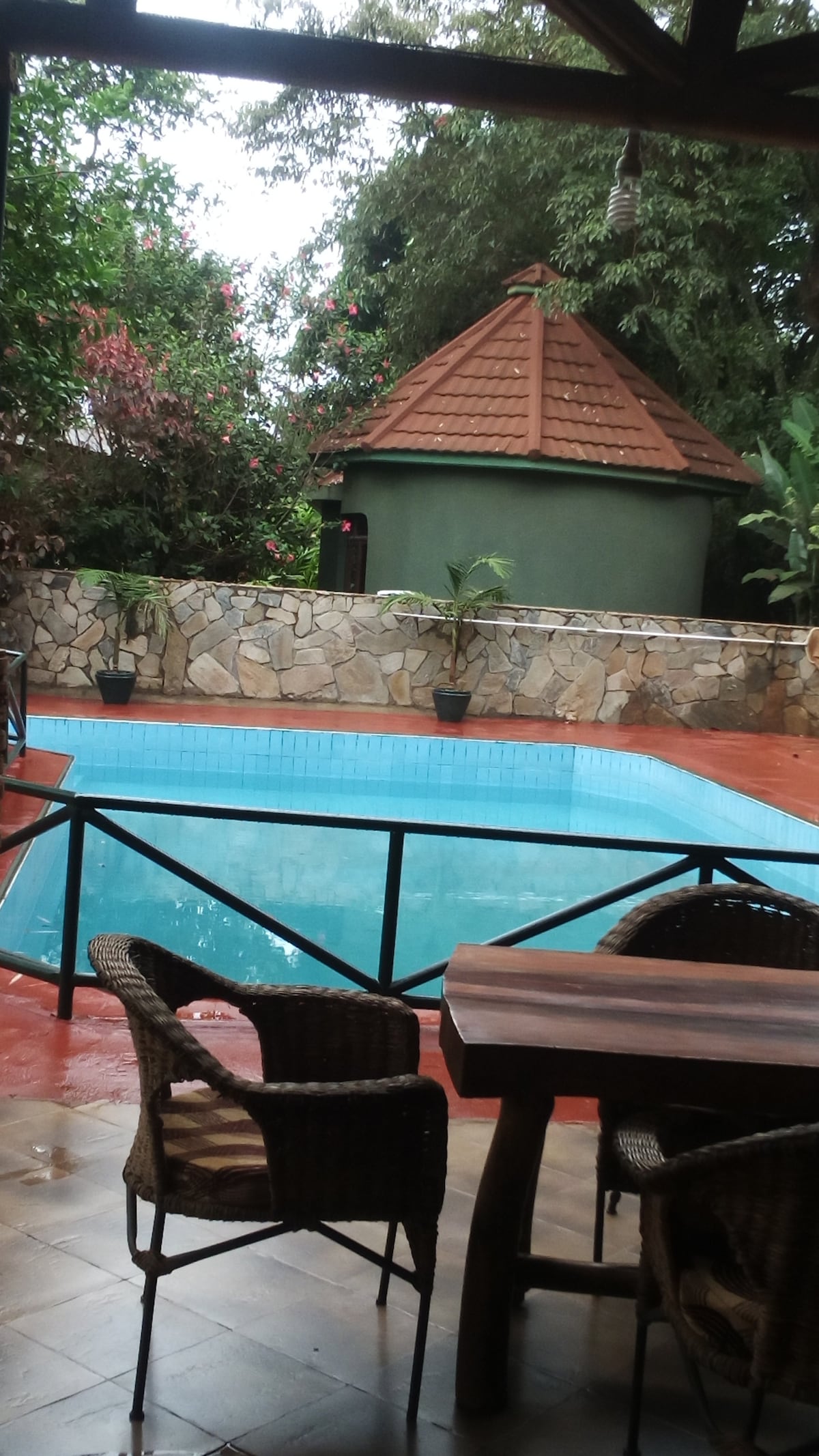 A swimming pool is framed by a wooden deck and surrounded by lush greenery. Nearby, a rustic dining set features woven chairs and a wooden table, offering a comfortable spot to relax. The backdrop includes a charming gazebo with a tiled roof.