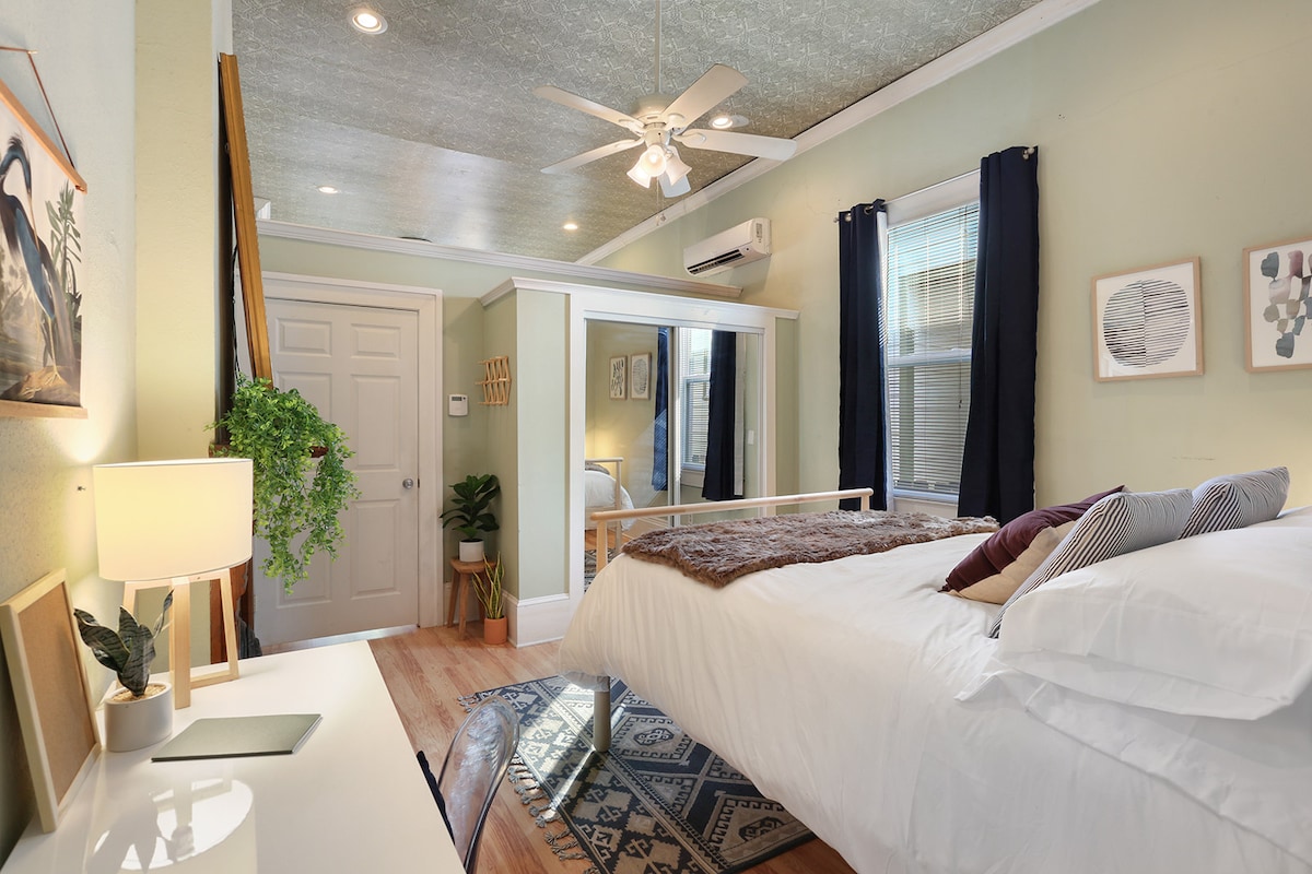 A bright bedroom features a spacious bed adorned with soft linens and decorative pillows. A desk with a lamp stands nearby, while windows dressed in dark curtains allow for natural light. Decorative wall art and a ceiling fan contribute to the inviting atmosphere.
