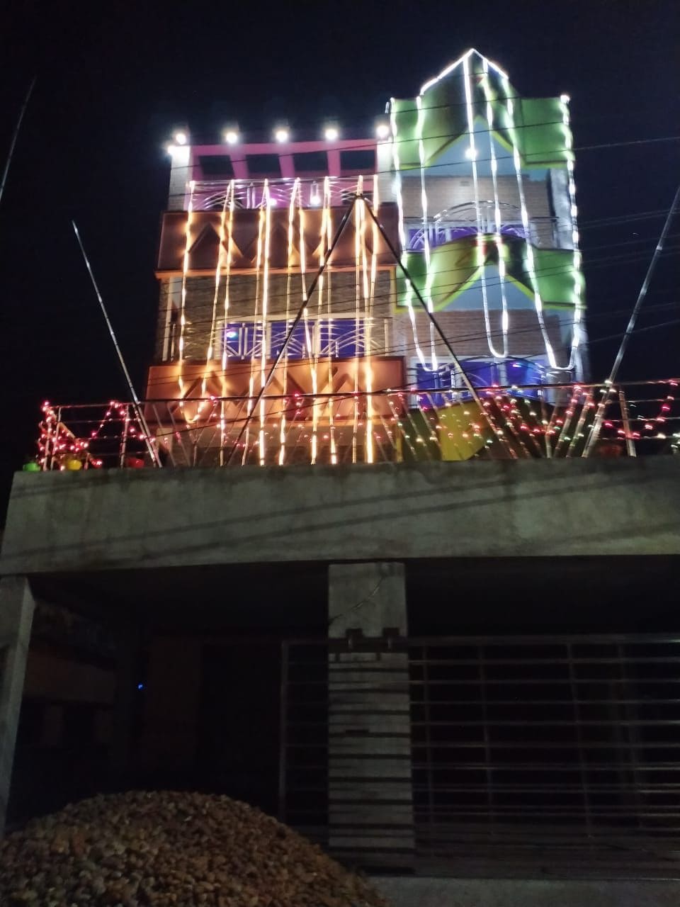 A modern multi-story building is illuminated with colorful lights, showcasing a vibrant facade. Decorative elements and artistic patterns create a lively appearance against the night sky. The structure sits above a stone-paved area, emphasizing its significant height and contemporary design.