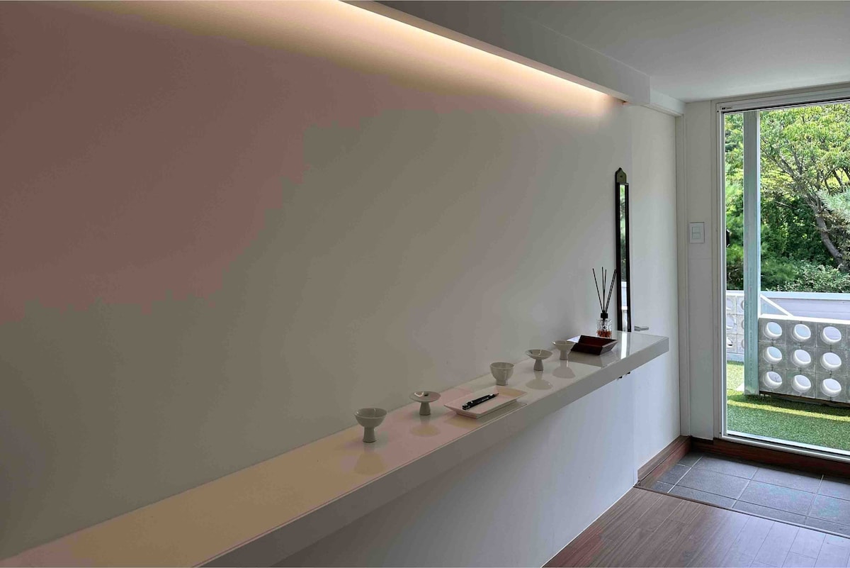 A minimalist interior space features a long white shelf adorned with ceramic items and an incense holder. Soft lighting illuminates the area, while a large window offers views of greenery outside, creating a calm and serene atmosphere.