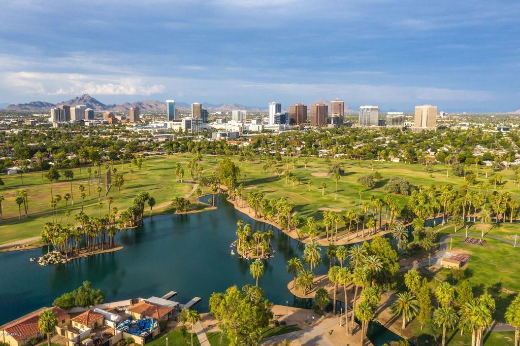 Aerial view showcases expansive green golf courses surrounded by palm trees and serene water features. The skyline of downtown Phoenix is visible in the background, highlighting a blend of urban and natural landscapes. Clear skies add to the picturesque scene.