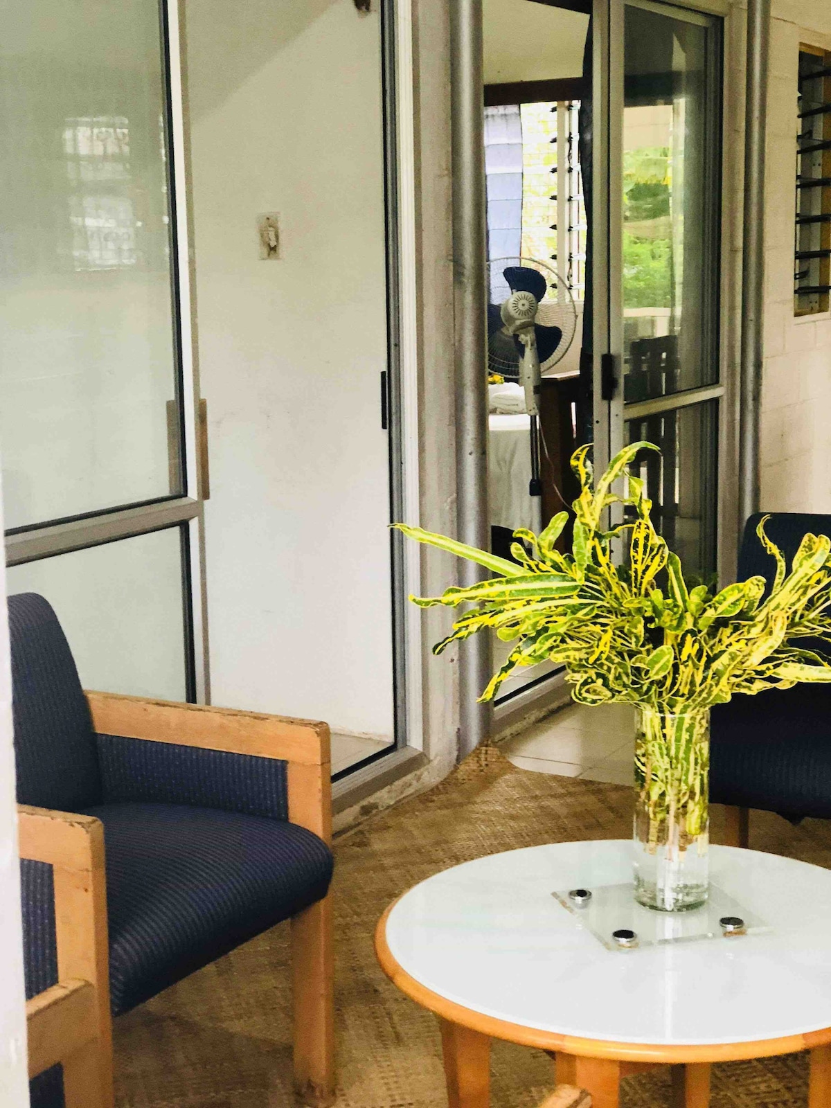 A cozy patio area is featured, showcasing two wooden chairs with navy fabric cushions. A circular glass table sits center stage, adorned with a vase of vibrant green foliage. Multiple sliding glass doors lead into the interior space, providing natural light.
