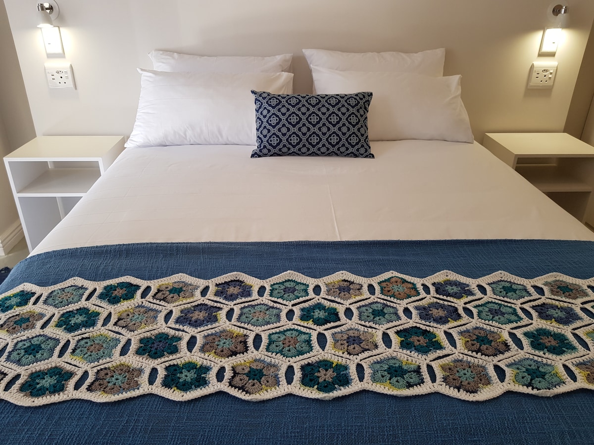 A queen size bed is adorned with crisp white linen and a colorful knitted throw layered across the duvet. Decorative pillows are placed centrally, while modern bedside tables are flanked on either side, each featuring subtle wall-mounted lighting.