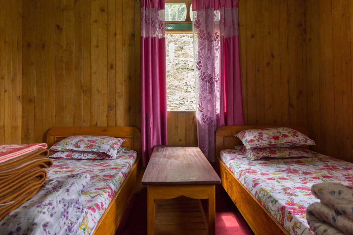 A cozy room featuring two twin beds adorned with floral-patterned bedding. In between the beds is a wooden table. Pink curtains frame the window, allowing natural light to illuminate the warm wooden walls.