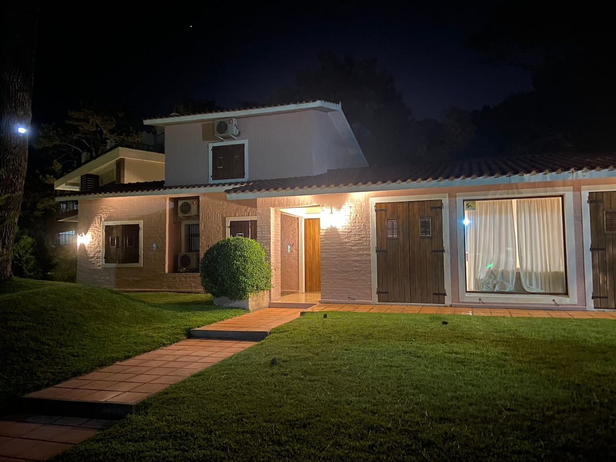 The exterior of a two-story house is illuminated at night, showcasing a well-maintained lawn and paths leading to the entrance. Warm lighting highlights the front door and windows, which feature light-colored curtains, creating a welcoming appearance against the dark sky.