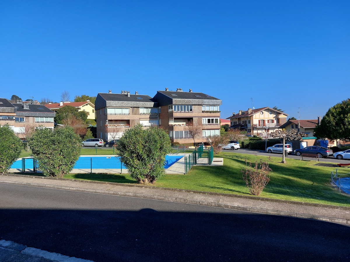 The image shows a peaceful urbanization with two stories of residential buildings. A swimming pool surrounded by grass and low greenery is visible in the foreground. Several parked cars line the street, contributing to the quiet residential atmosphere.