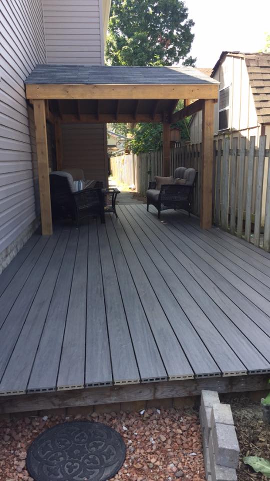 A covered outdoor seating area is featured, with two comfortable chairs positioned on a wooden deck. The space is flanked by a fence, providing privacy and a sense of seclusion. Natural light filters through the surrounding greenery.