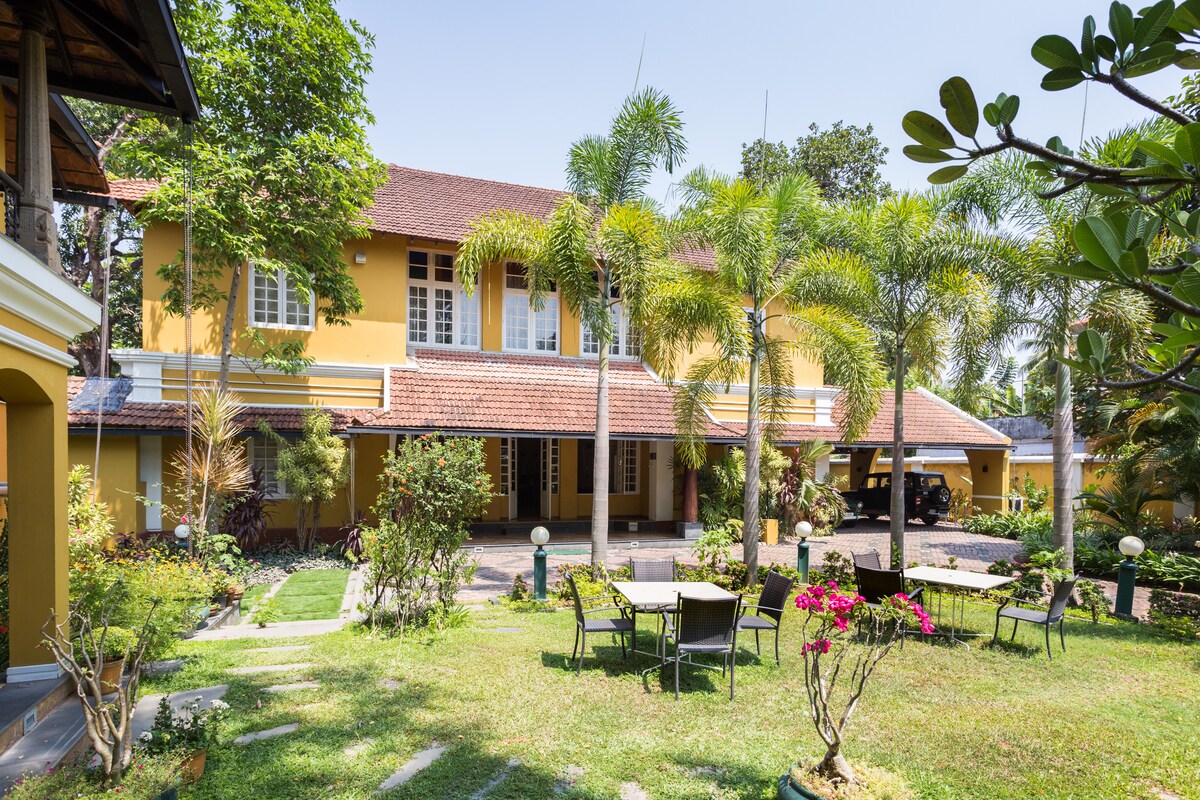 A charming outdoor space is featured, showcasing a well-kept garden with manicured lawns and colorful flower beds. The two-story colonial-style building stands in the background, framed by palm trees, while several chairs and tables invite relaxation in the sunshine.