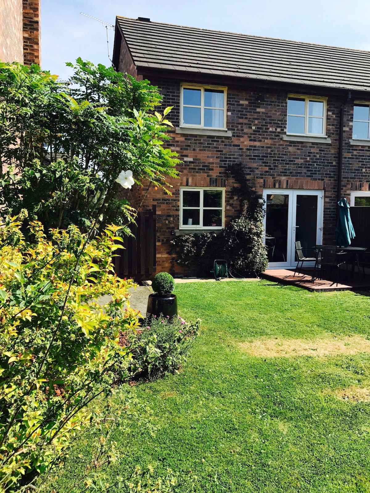 The well-maintained garden is shown, featuring a lush green lawn bordered by colorful shrubs and plants. The house is seen in the background, with French doors leading to the outdoor sitting area. A patio is visible, complemented by outdoor furniture, creating a spacious environment.