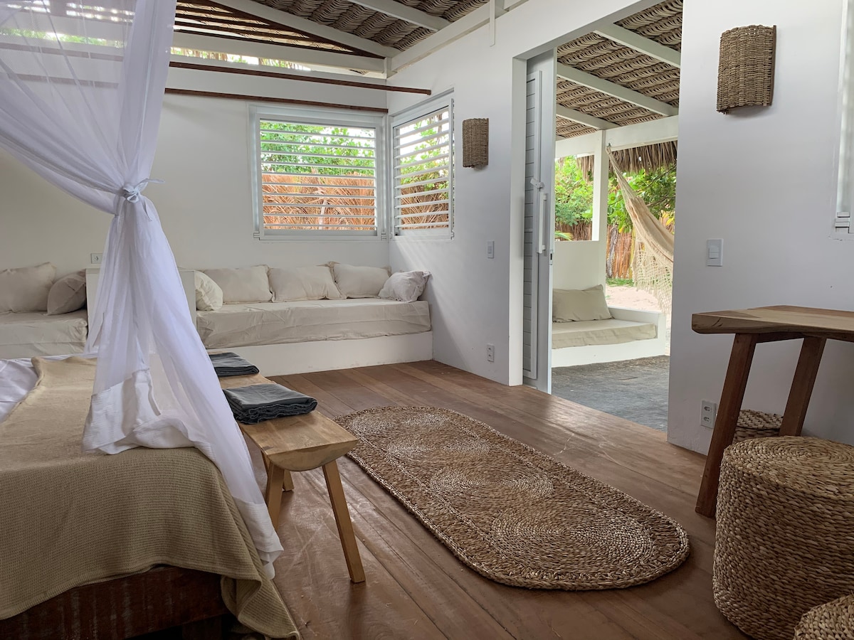 A bright and airy chalet interior features a spacious bed draped with sheer fabric. Cozy seating areas with plush cushions are arranged by the windows, and a natural fiber rug lies on the wooden floor. A wooden table and stools offer additional seating options.